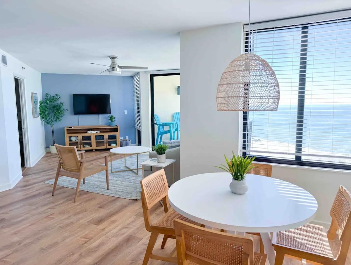 A bright dining area features a round white table surrounded by four wooden chairs. A plant is centered on the table. In the background, a living area is visible, showcasing a wall-mounted television and seating arrangement. Large windows allow for natural light and showcase views of the Gulf.