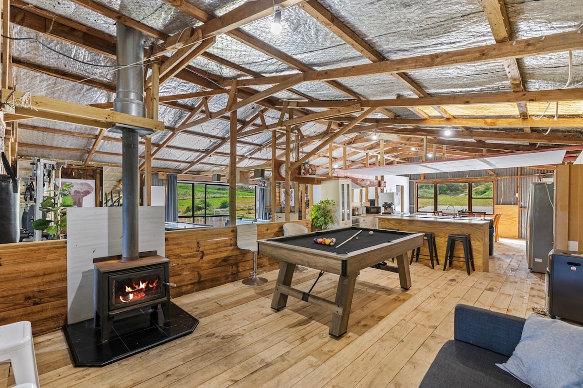 An open-plan living space is showcased with wooden floors and a high exposed beam ceiling. A pool table occupies the center, complemented by a cozy wood-burning stove. Large windows allow natural light to fill the room while offering views of the surrounding landscape.