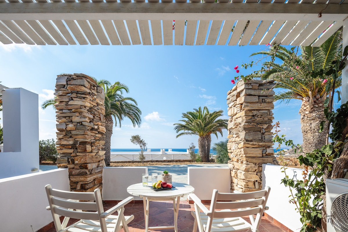 An outdoor seating area features a round table accompanied by two white chairs. Stone columns flank the space, providing support for a wooden canopy. Palm trees and the sea are visible in the distance, enhancing the serene environment.