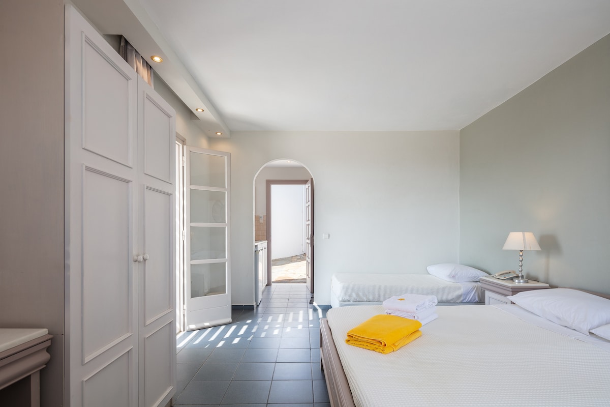 A spacious guest room featuring two beds and ample natural light from a glass door leading to an outdoor area. A bedside table with a lamp is positioned next to one bed. Fresh white towels and a bright yellow bath towel are neatly arranged on the bed.