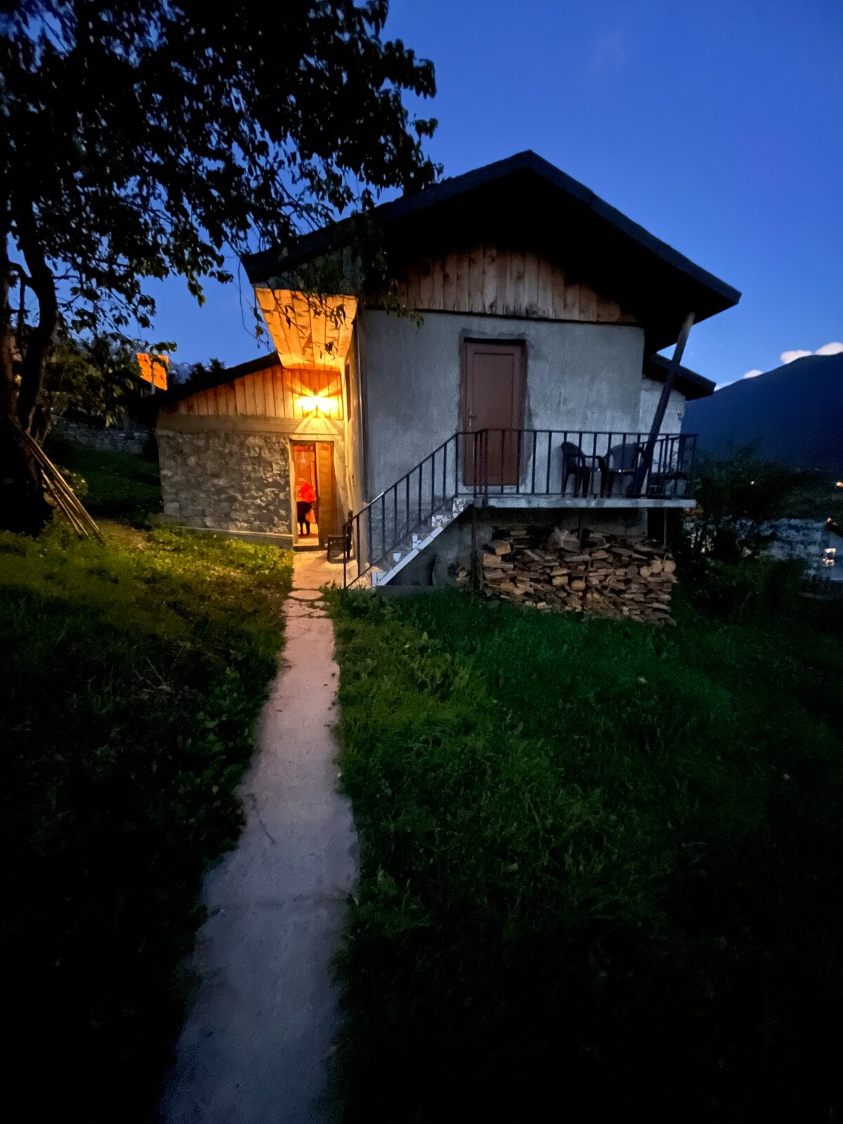 A charming two-story building is positioned on a grassy slope, with a stone pathway leading to the entrance. Soft light emanates from an illuminated doorway, creating a welcoming ambiance. Stacked firewood is neatly arranged beside the entrance, enhancing the rustic charm.