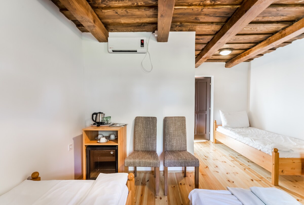 A bright, simple sleeping area features two single beds with white linens. A small kitchenette corner with a kettle is visible, alongside two upholstered chairs placed near a wooden table. The room showcases wooden beams across the ceiling and light-colored wooden flooring.