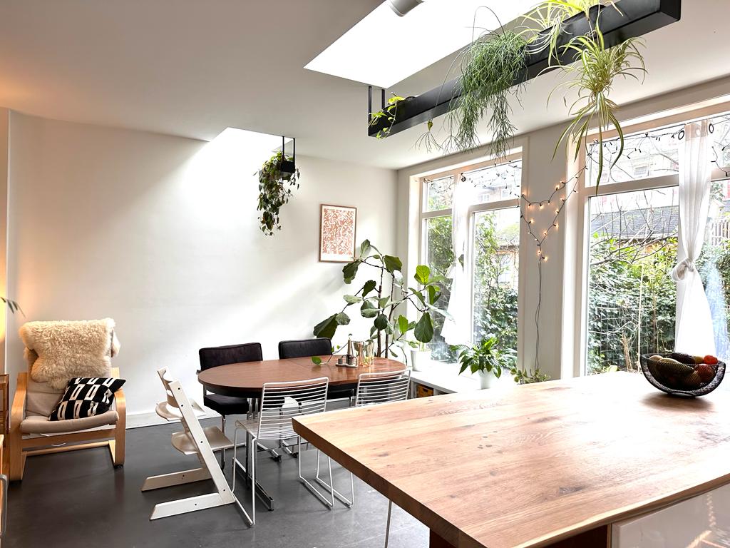 A bright dining area is showcased with large windows promoting natural light. A circular wooden table is surrounded by white chairs, while plants hang from the ceiling and nearby surfaces. A cozy chair is positioned in the corner, creating a relaxed seating atmosphere.