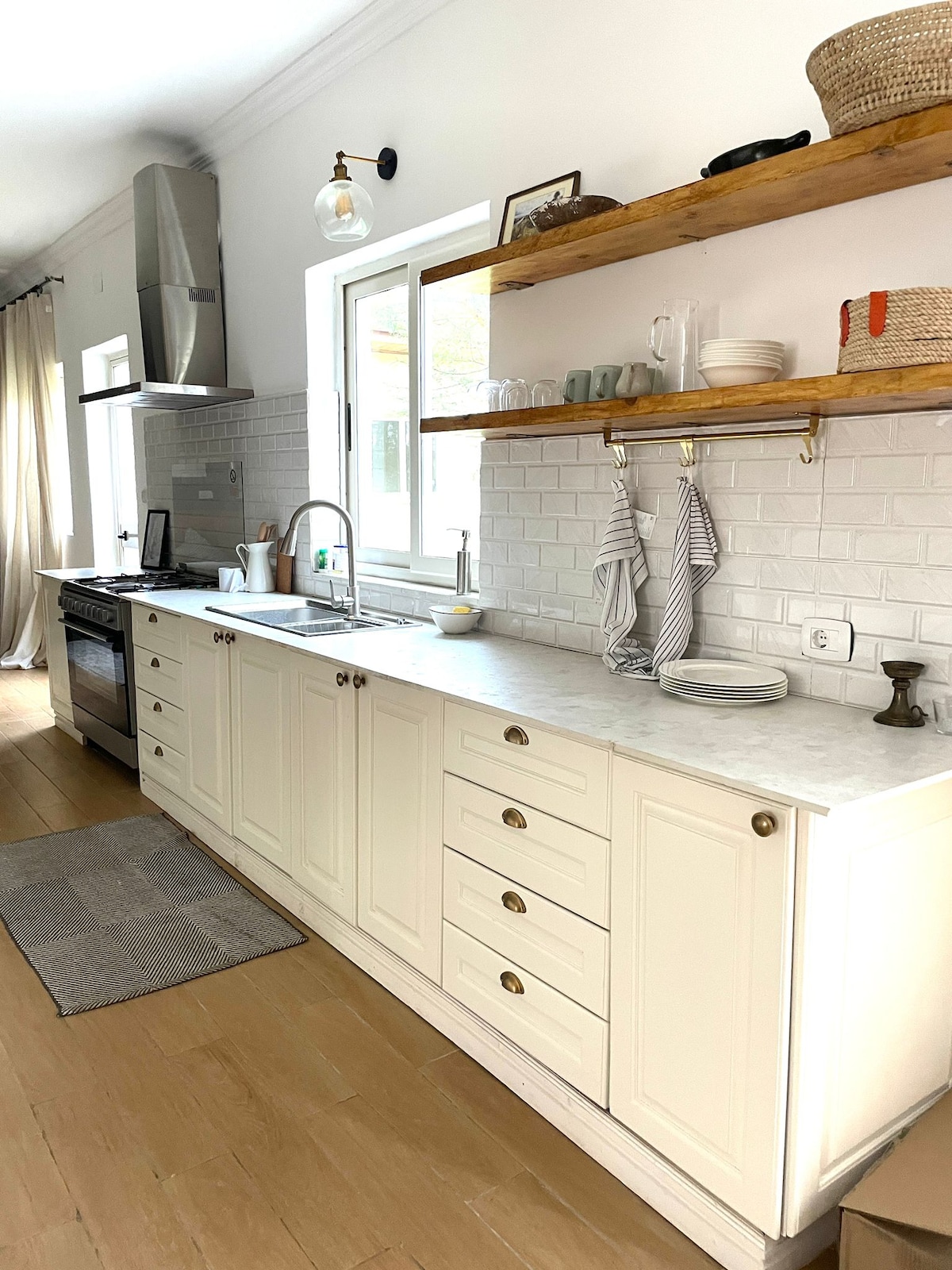 A bright kitchen features a clean marble countertop with ample cabinet space below. Open shelving displays glassware and decorative items. A modern stove and oven are installed, complemented by a sleek stainless steel range hood. Soft light enters through the window, enhancing the inviting space.