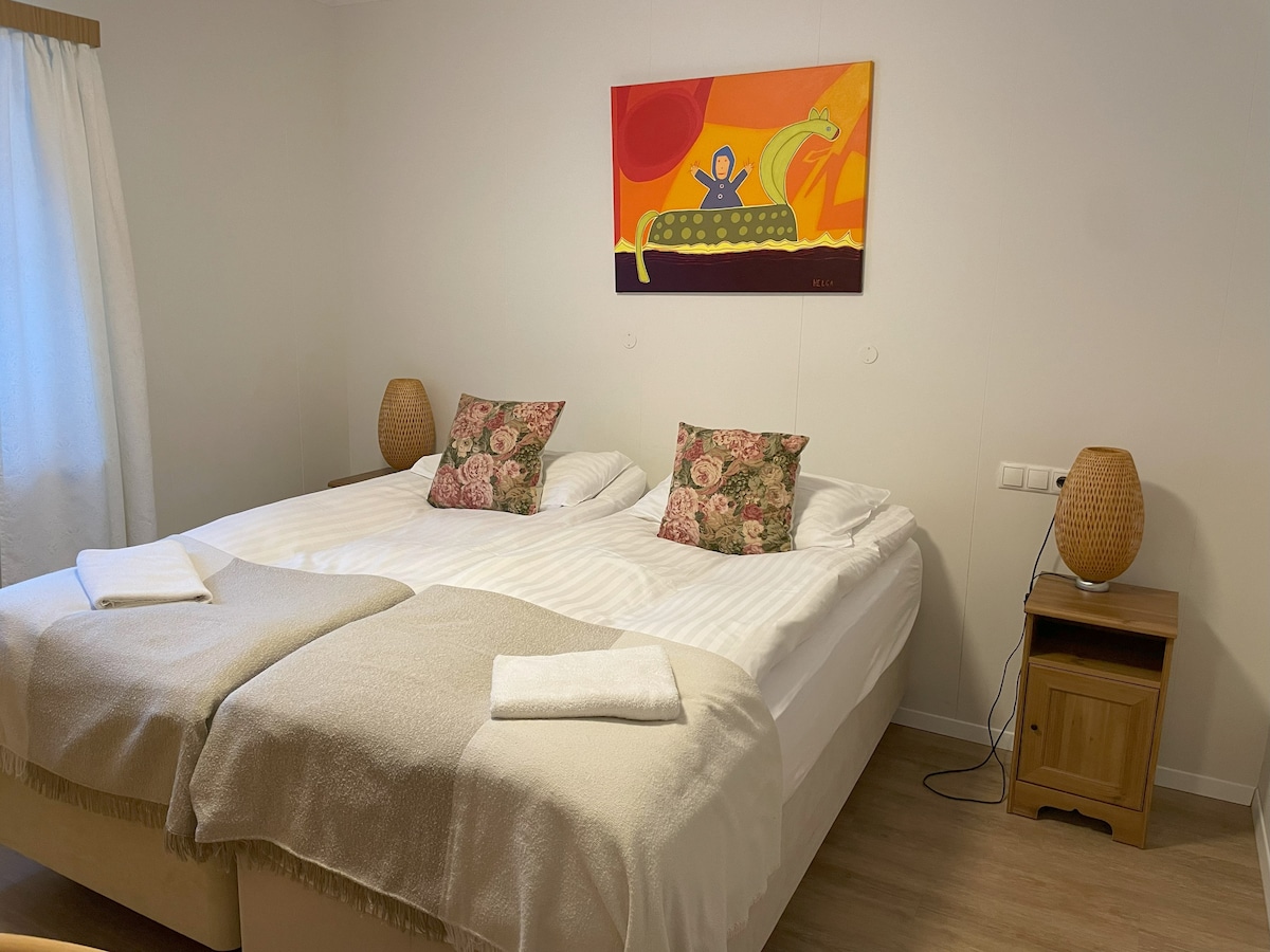 The sleeping area features two spacious beds, each adorned with white linens and a decorative throw. Colorful wall art adds a playful touch, while bedside lamps provide gentle illumination. Natural light filters through a curtain, enhancing the inviting atmosphere of the room.