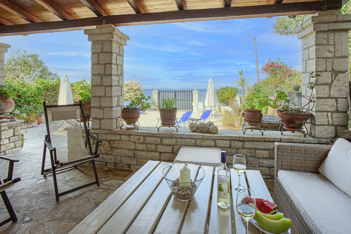 A shaded outdoor seating area features a wooden table set with a glass bowl and beverages. Comfortable chairs surround the table, while potted plants and sun loungers are visible in the background, offering a view of the sea and garden landscape.