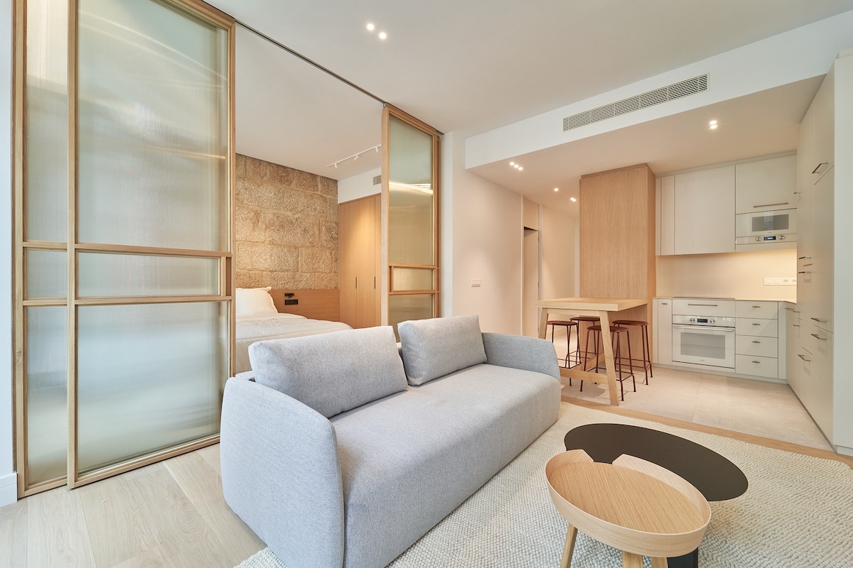 An open living area is observed, featuring a light grey sofa paired with a round coffee table. Glass-paneled partitions separate the sleeping area, which includes a neatly made bed. A modern kitchenette with wooden accents and four stools is also visible in the background.
