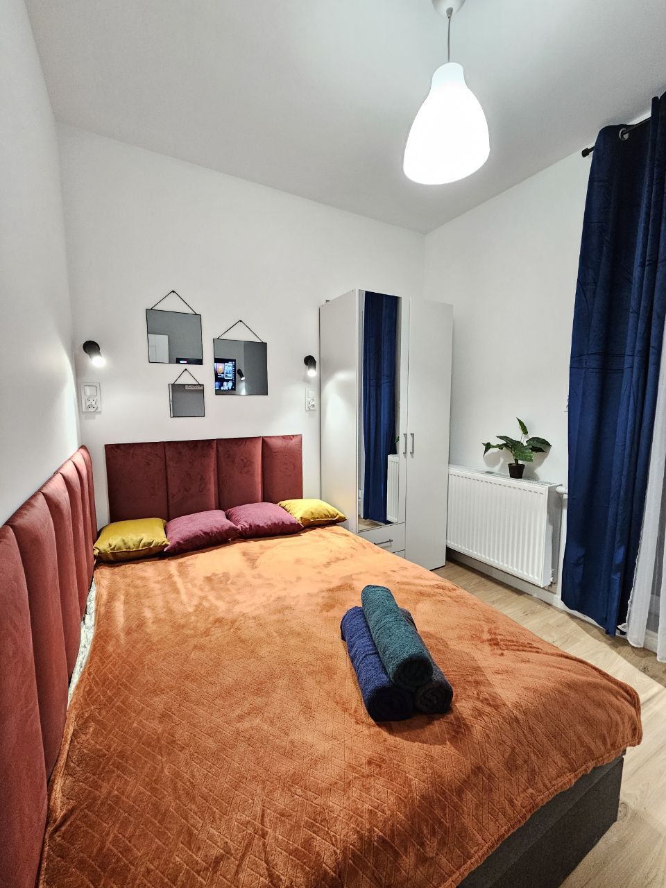 A cozy sleeping area features a spacious bed dressed in a vibrant orange blanket. Two accent pillows are placed at the head of the bed. A wardrobe stands nearby, and a plant adds a touch of greenery. Decorative mirrors are mounted on the wall, reflecting light.