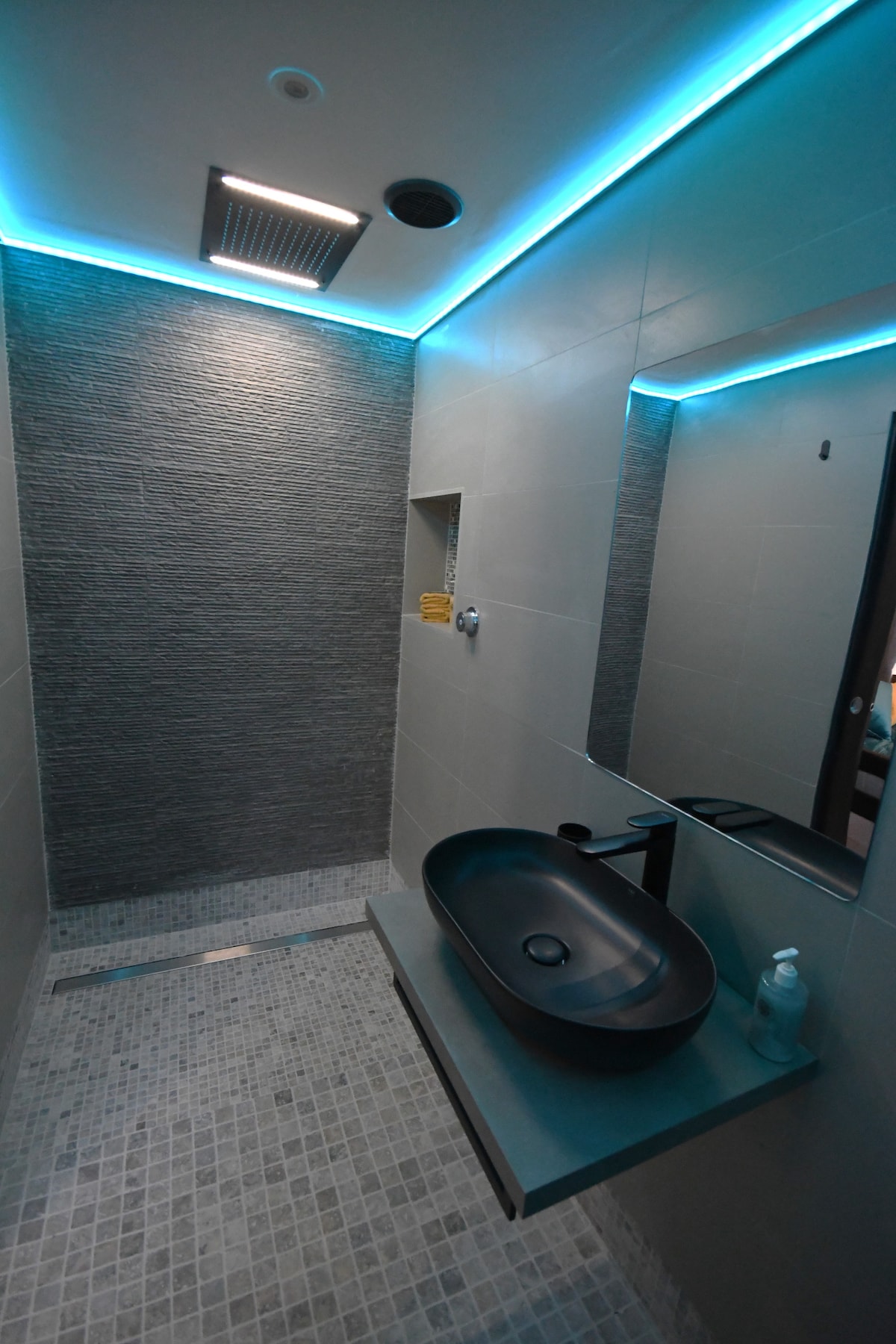 The ensuite wet room features contemporary design with textured gray walls and a spacious shower area. A sleek black basin is positioned on a floating vanity, complemented by a large mirror above. Illuminated edges provide soft lighting, enhancing the modern aesthetic.