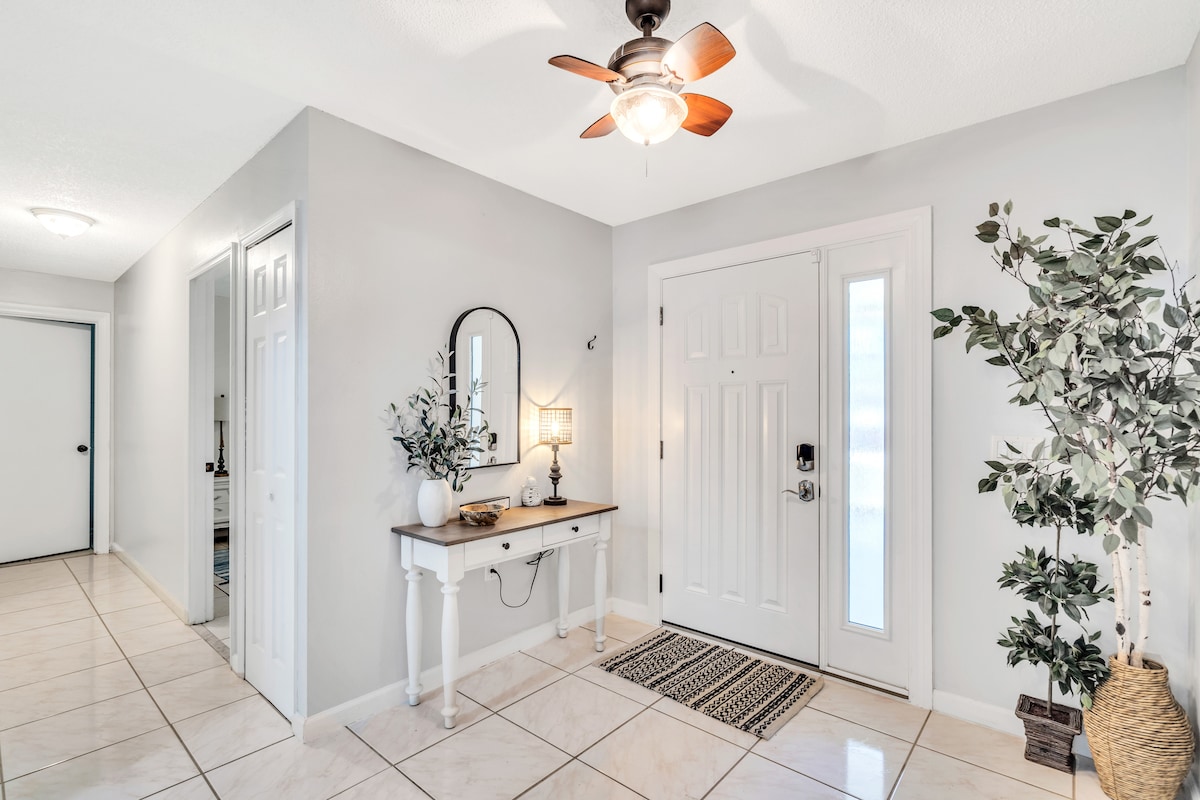 A welcoming entryway features a white console table with decorative plants and a round mirror. A ceiling fan provides air circulation. The tile flooring and light-colored walls create a bright atmosphere, leading into the rest of the home.