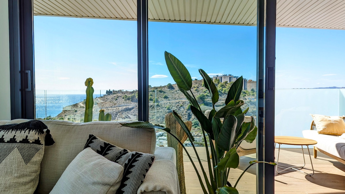 A bright interior space features large glass doors that reveal a stunning ocean view. A comfortable sofa is adorned with patterned cushions, while potted plants add a touch of greenery. Natural light floods the area, enhancing the connection to the outdoor scenery.