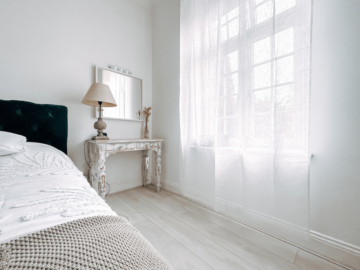A bright bedroom features a spacious bed with white linens, complemented by a textured throw. A light wood floor enhances the airy feel. A vintage-style lamp stands on a decorative bedside table next to a large window draped with sheer curtains, allowing soft natural light to fill the room.