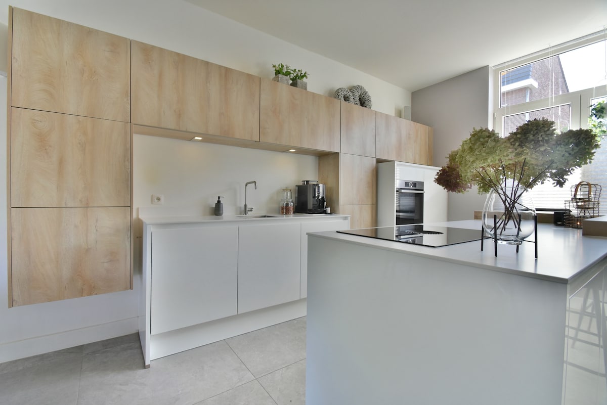 A modern kitchen area features sleek white cabinets complemented by natural wood accents. A stainless steel oven and modern appliances are integrated into the design. Natural light fills the space through a large window, with decorative greenery providing a touch of freshness on the counter.