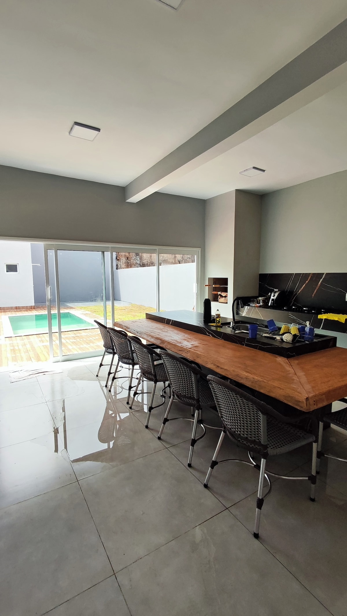 A spacious kitchen area is equipped with a large wooden countertop and six metal chairs. Natural light flows in through sliding glass doors that open to an outdoor space featuring a pool and a grassy area.