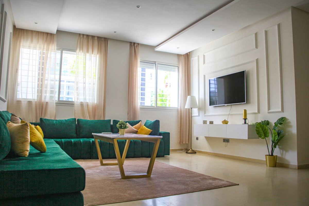 A bright living room features a spacious, L-shaped green sofa adorned with cushions, positioned around a stylish coffee table. Natural light filters through large windows, illuminating the elegant decor. A wall-mounted TV is visible, enhancing the room's modern design.