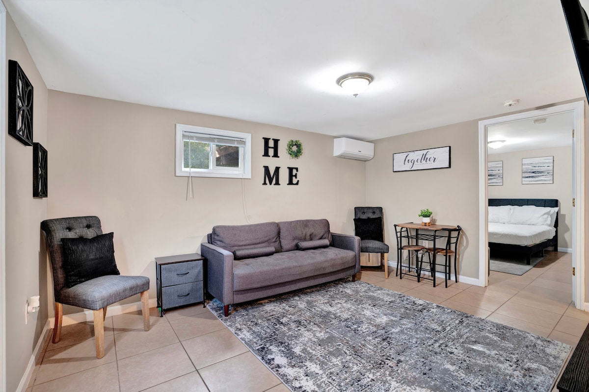 A cozy living area is presented with a gray sofa, two armchairs, and a small dining table with two chairs. Wall decor features the word 'HOME' and minimalist artwork. Natural light enters through a window, illuminating the space. The entrance to a bedroom is visible in the background.