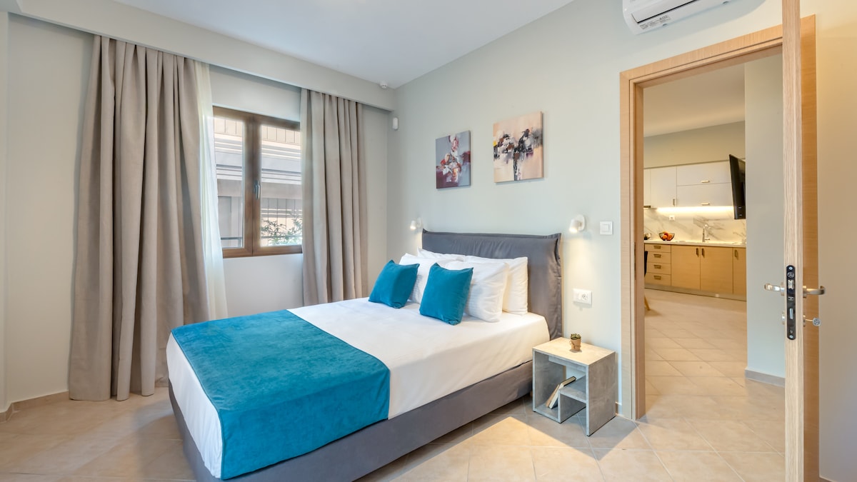 A modern bedroom features a spacious bed dressed in crisp white linens and a teal throw. Two framed artworks hang above the bed, with soft lighting provided by wall-mounted lamps. Natural light filters through a large window adorned with light curtains.
