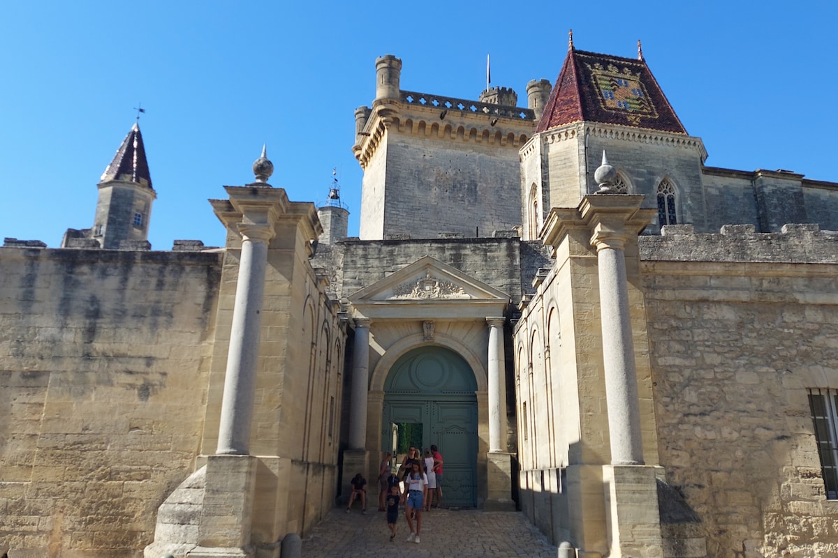 An imposing historical building is displayed, featuring stone walls and a large archway entrance. Towering turrets are visible, adorned with distinctive roofing. The scene includes several visitors walking toward the entrance, emphasizing the scale and grandeur of the structure against the clear blue sky.