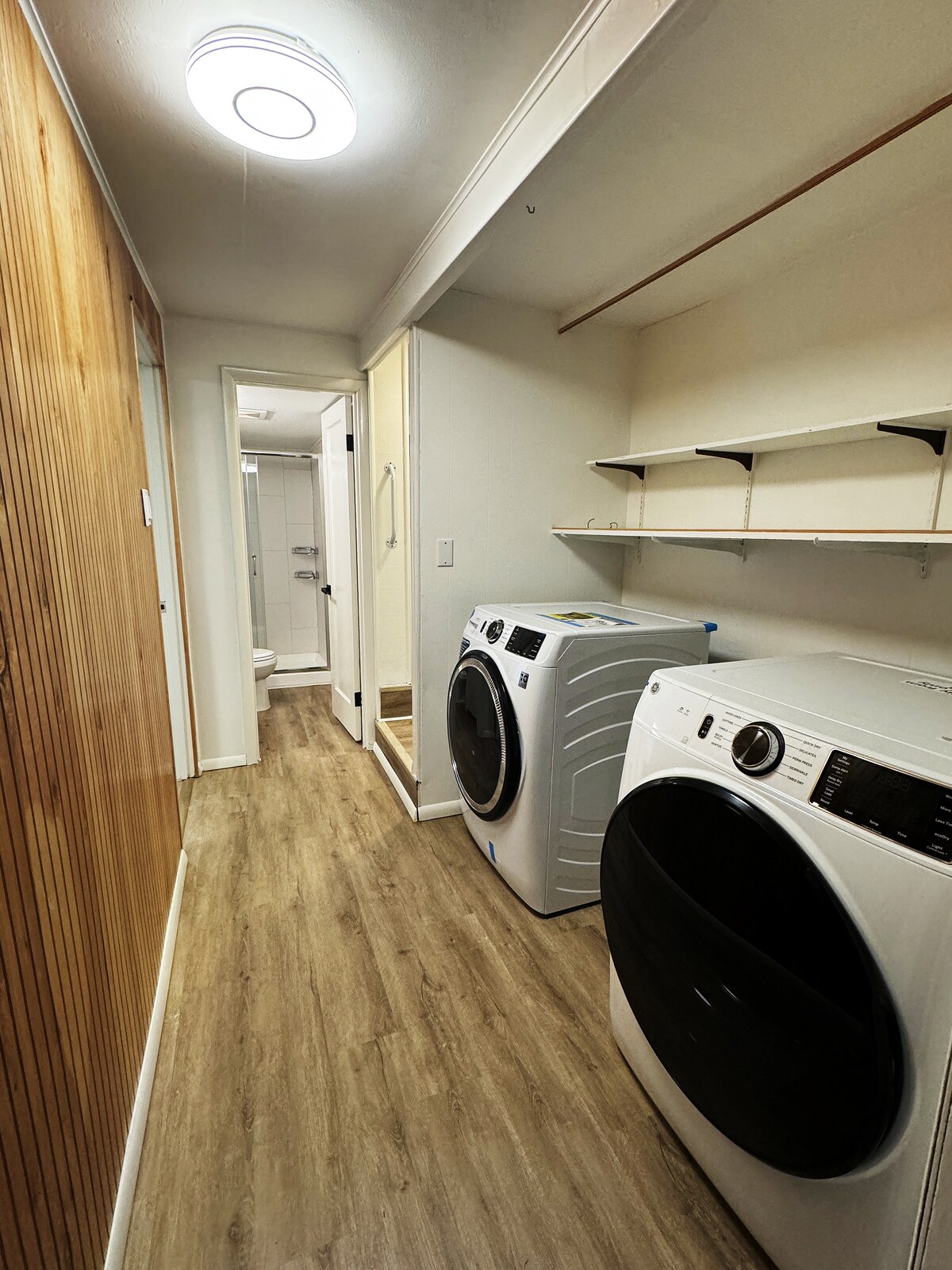 A designated laundry space is equipped with a washer and dryer, positioned along a neutral-toned wall. Shelving is available overhead for storage. In the background, a door leads to a bathroom, and light-colored flooring lends a clean and functional atmosphere.