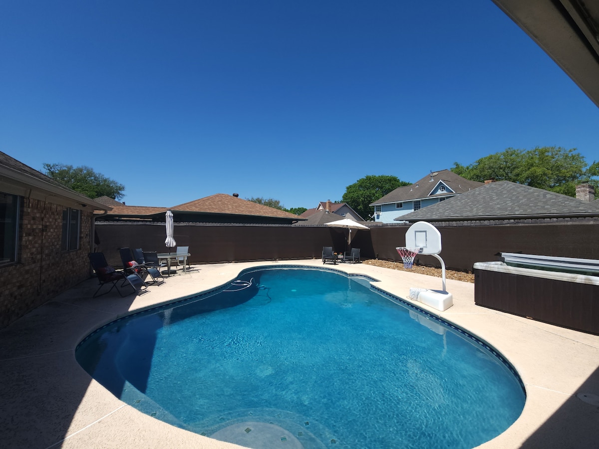 A large swimming pool with a rounded shape reflects a clear blue sky. Poolside seating is arranged with several lounge chairs, and a basketball hoop is positioned at one end. A hot tub is located nearby, surrounded by a privacy fence.
