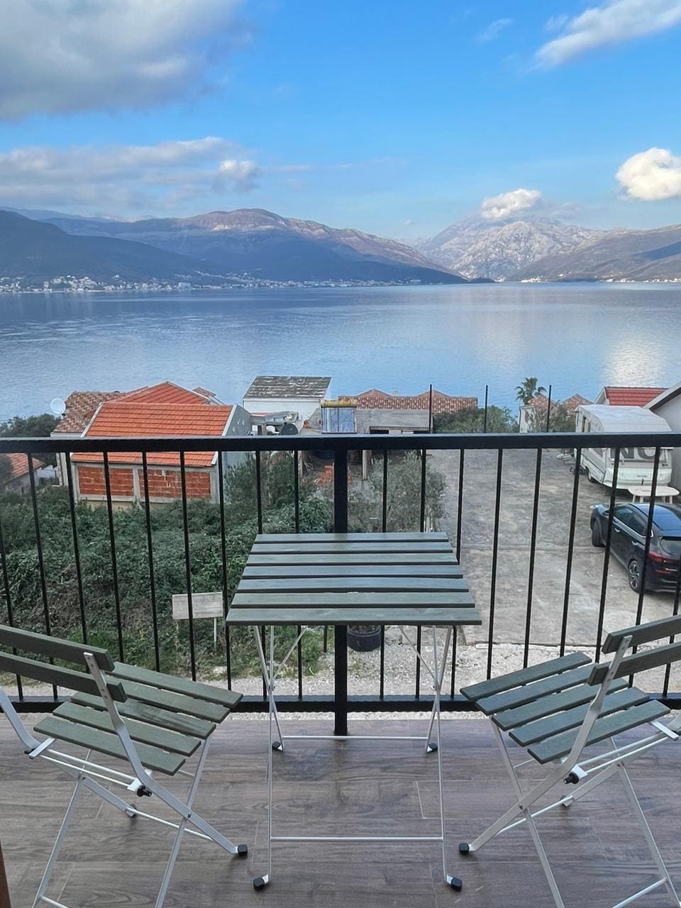 A small balcony is shown with two foldable chairs and a table, overlooking the tranquil waters of Boka bay. The scenery features distant mountains and a glimpse of nearby rooftops under a partly cloudy sky.