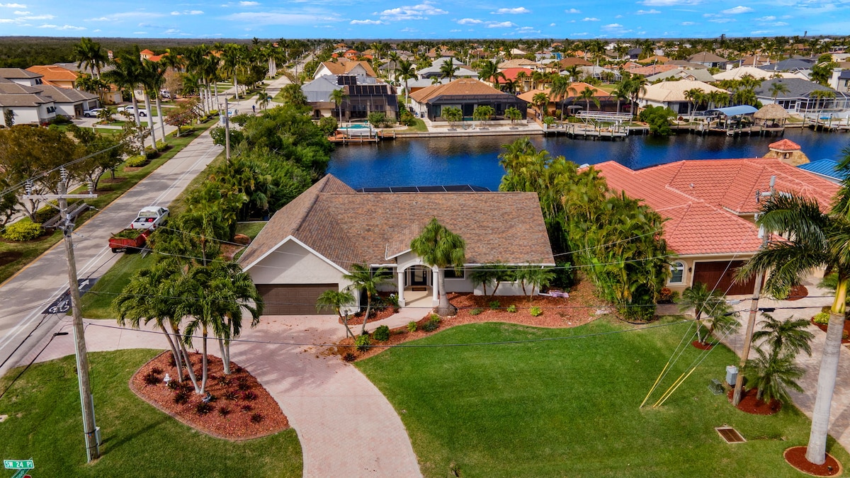 This aerial view showcases a single-family home set on a spacious lot with a well-maintained lawn. Surrounding palm trees and tropical landscaping contribute to the outdoor ambience. A serene canal adjacent to the property reflects the neighborhood's charm, featuring various homes in the background.