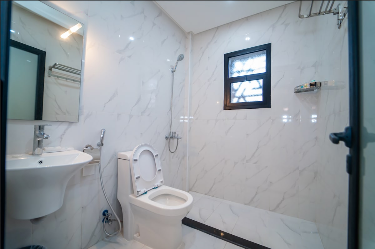 A modern bathroom is presented with light marble walls and a sleek, clean design. A showerhead is mounted above, and a small shelf for toiletries is attached to the wall. Natural light filters in through a window, enhancing the bright atmosphere.