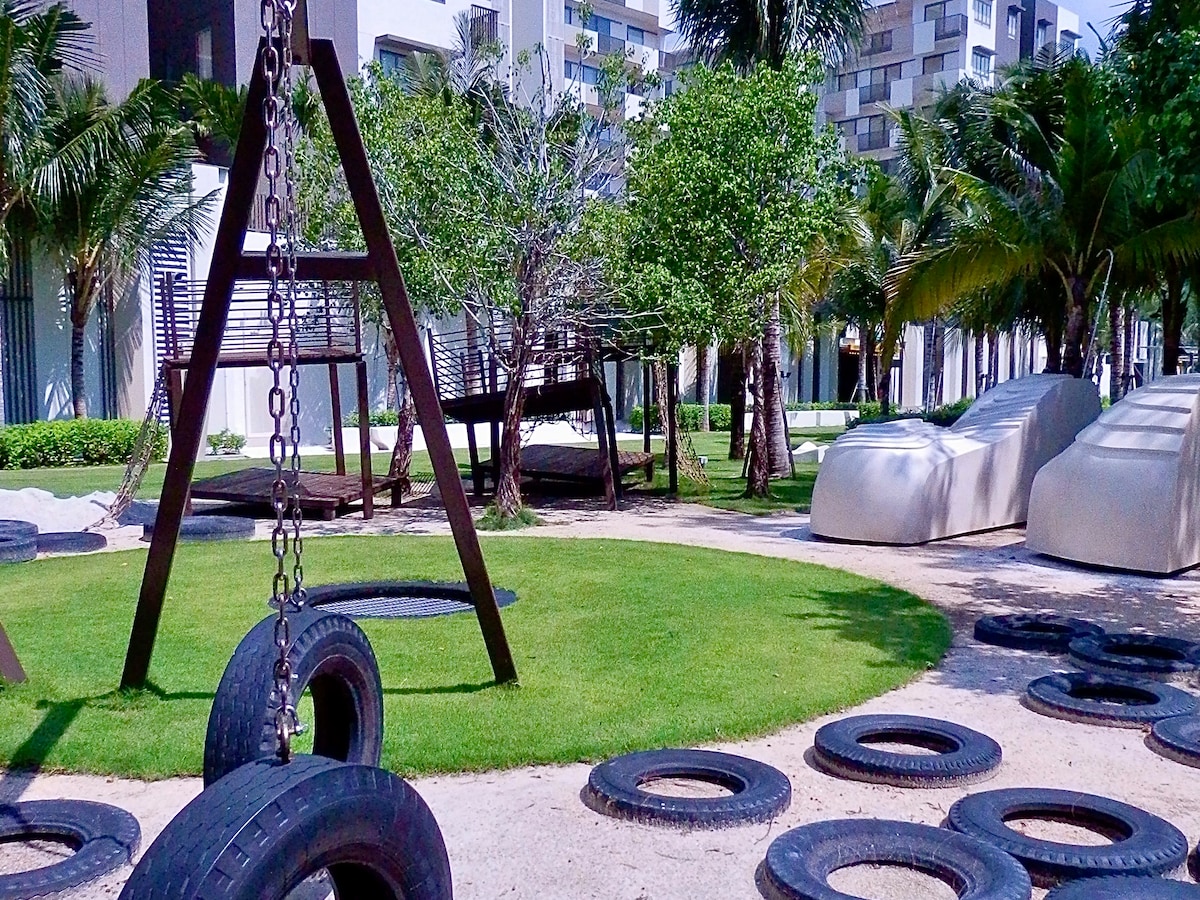 An outdoor playground area features a swing set with a tire swing and a grassy space surrounded by palm trees. A wooden climbing structure with ropes is positioned nearby, along with distinct molded seating areas. The atmosphere exudes fun and engagement for children.