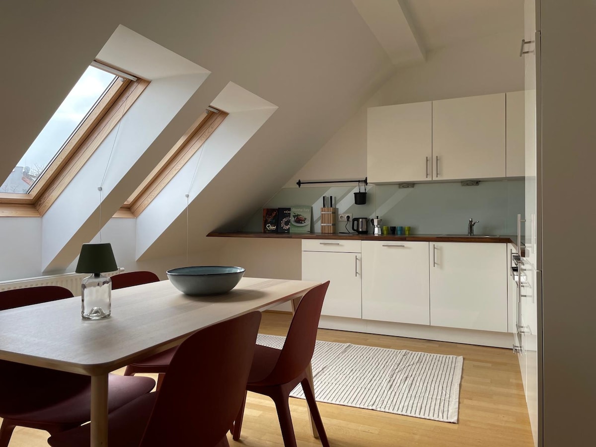 A bright kitchen area with sloped ceilings features modern cabinetry and appliances. A wooden dining table is surrounded by four red chairs. Large windows allow natural light to fill the space, highlighting a bowl and a decorative table lamp on the table.