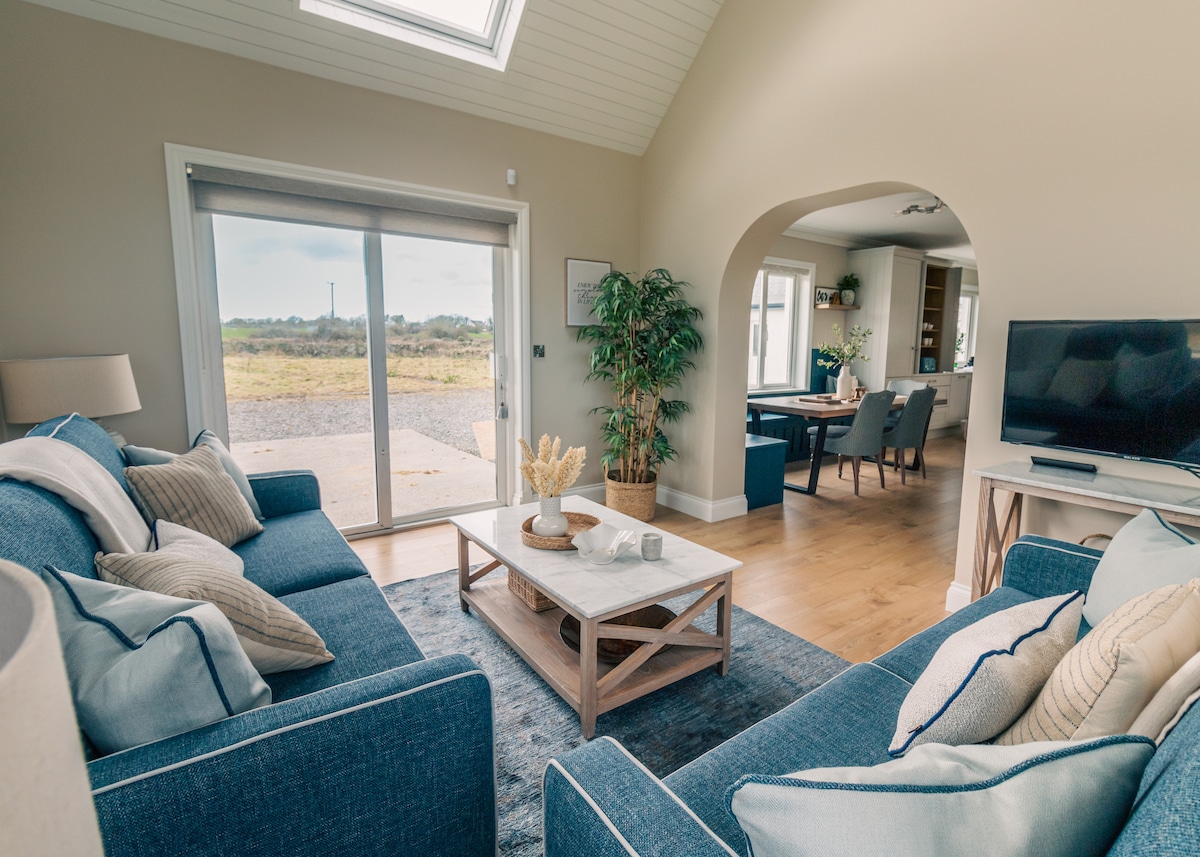 A spacious living area is presented, featuring two blue sofas adorned with decorative pillows. A light wooden coffee table rests on a soft area rug. Bright natural light pours in through the large sliding doors, which open to scenic outdoor views.