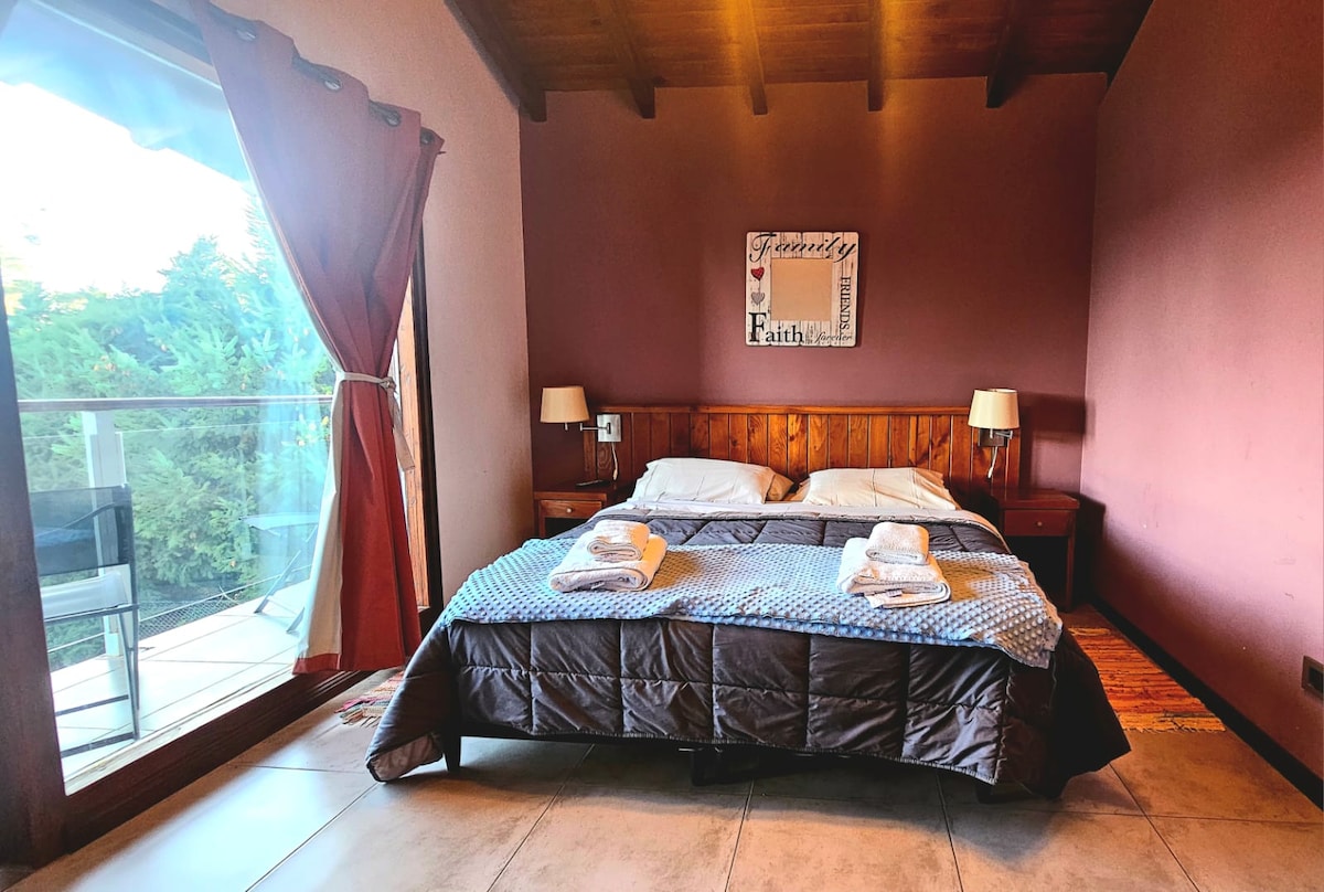 A spacious sleeping area features a large bed, adorned with neatly arranged towels and a cozy quilt. Two bedside tables with lamps are positioned alongside. Natural light filters in through the glass door, which opens to a balcony overlooking greenery.