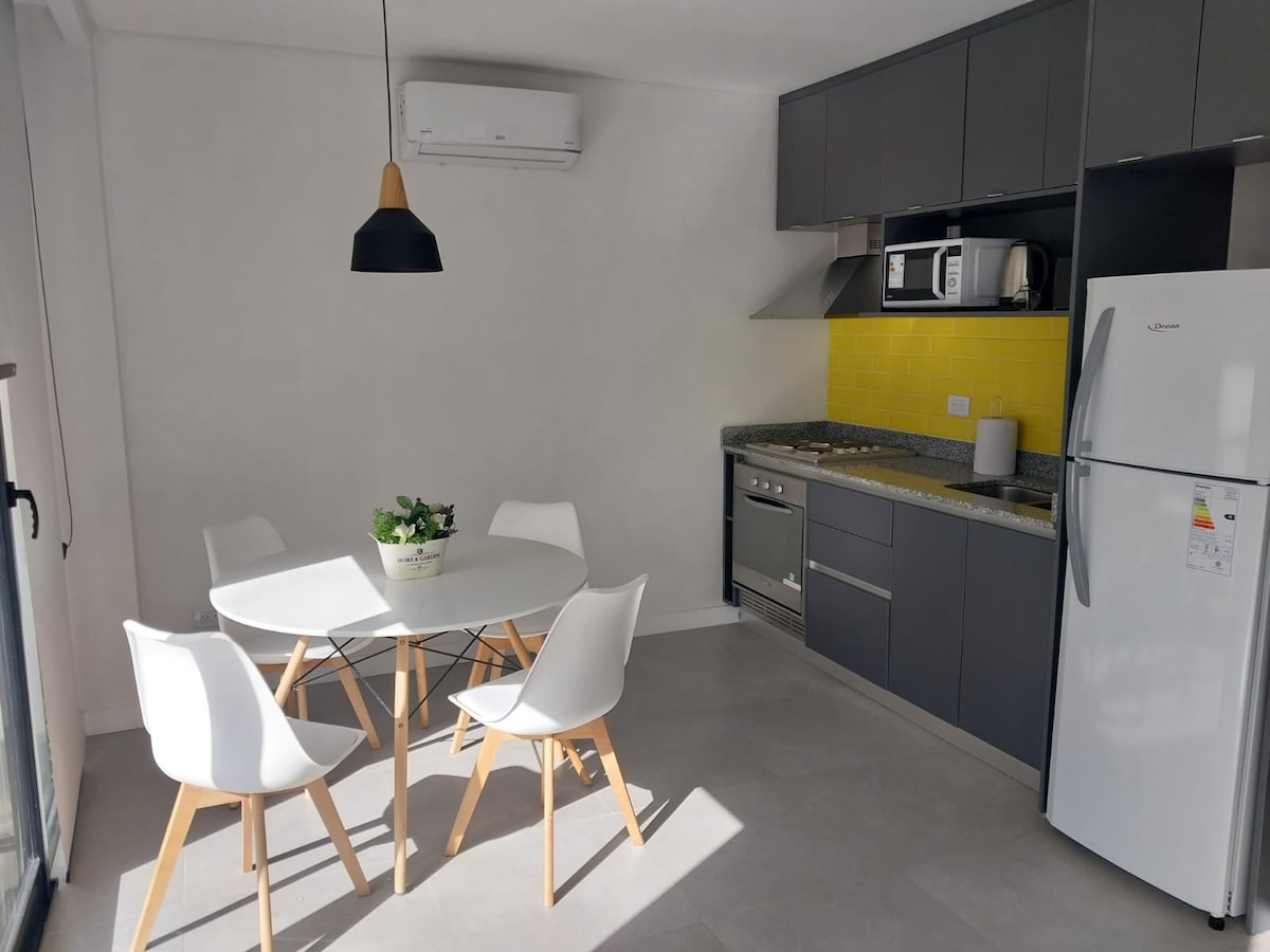 A modern kitchen area features sleek gray cabinets and a bright yellow backsplash. A round dining table with four white chairs is positioned beside the kitchen counter. A refrigerator, microwave, and oven are visible, with natural light filtering in through a large window.