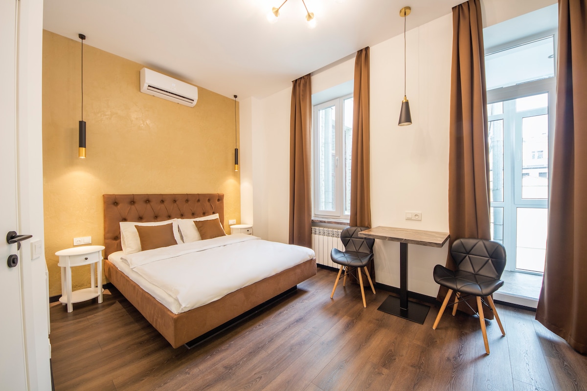 A spacious sleeping area features a large bed with neutral bedding against a textured accent wall. Two stylish chairs and a small table are positioned near the window, which is framed by floor-length curtains. Natural light fills the room from the window and double doors.