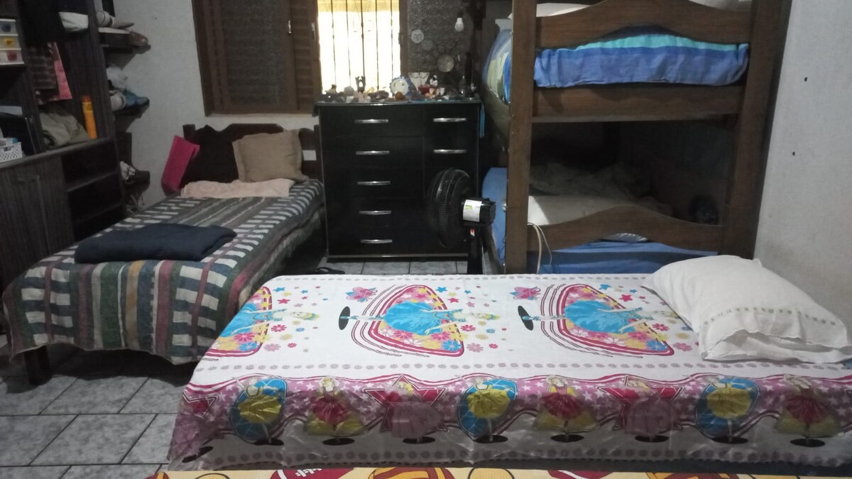 The room features a combination of bedding arrangements, including a spacious bed, a single bed, and a bunk bed in the background. A dark wooden dresser is positioned against the wall, adding storage space. Decorative quilts and pillows enhance the cozy atmosphere.