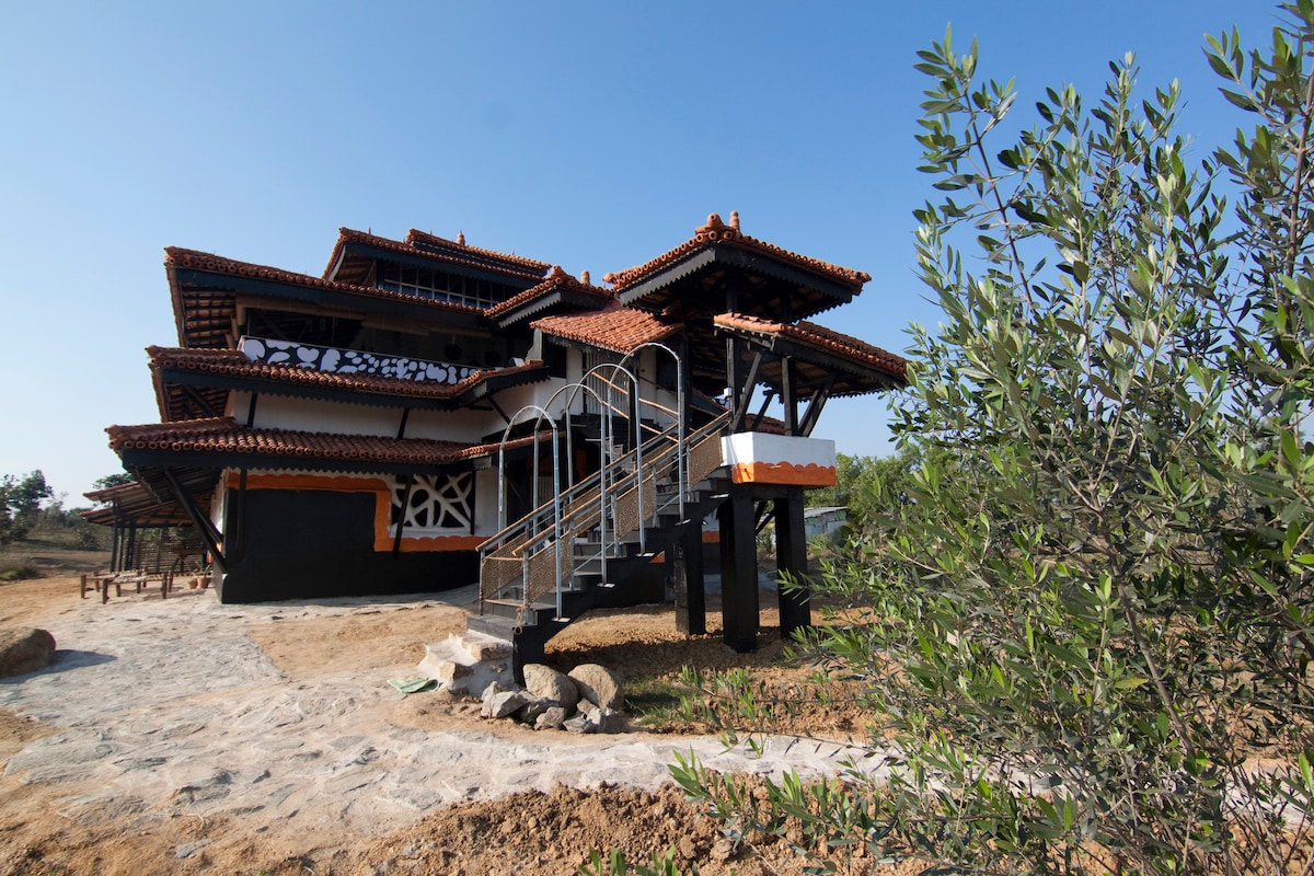 The exterior of the mud house is showcased, featuring traditional architecture with a distinctive roofline. Stairs lead to the entrance, bordered by natural landscaping that harmonizes with the rustic surroundings. The structure is complemented by a clear blue sky overhead.