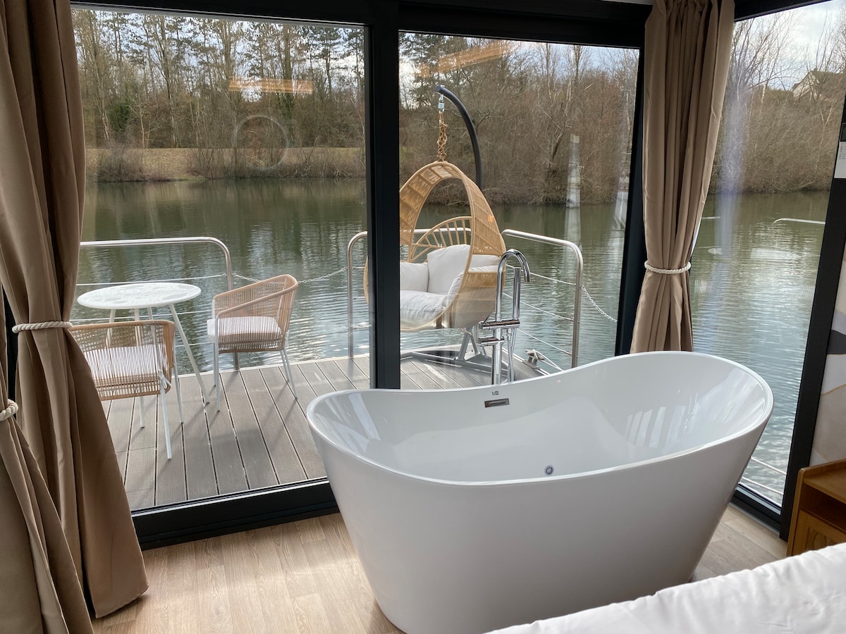 A sleek, freestanding bathtub is positioned beside large glass windows offering views of the tranquil water. Outside, a suspended chair and a small seating area provide a serene spot to enjoy the surrounding nature. Soft curtains frame the scene, adding a touch of elegance.