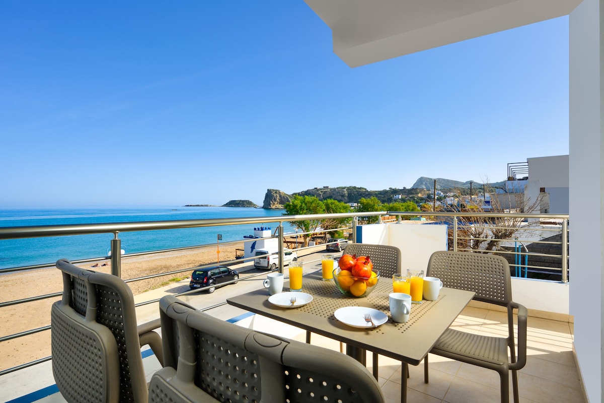 A balcony offers a dining table set with plates, glasses, and a fruit bowl. The view features sandy beach and the serene blue waters, framed by distant green hills under a clear sky. Comfortable seating surrounds the table, inviting relaxation.