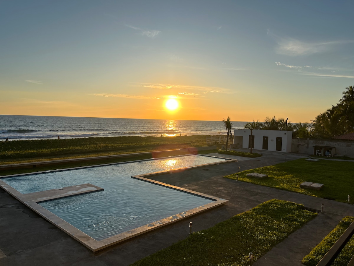 A tranquil scene captures the sun setting over the ocean, reflecting warm hues across the water. The shared swimming pool is viewed from a distance, surrounded by lush green grass and palm trees. A spacious area with lounge chairs can be seen alongside the pool.