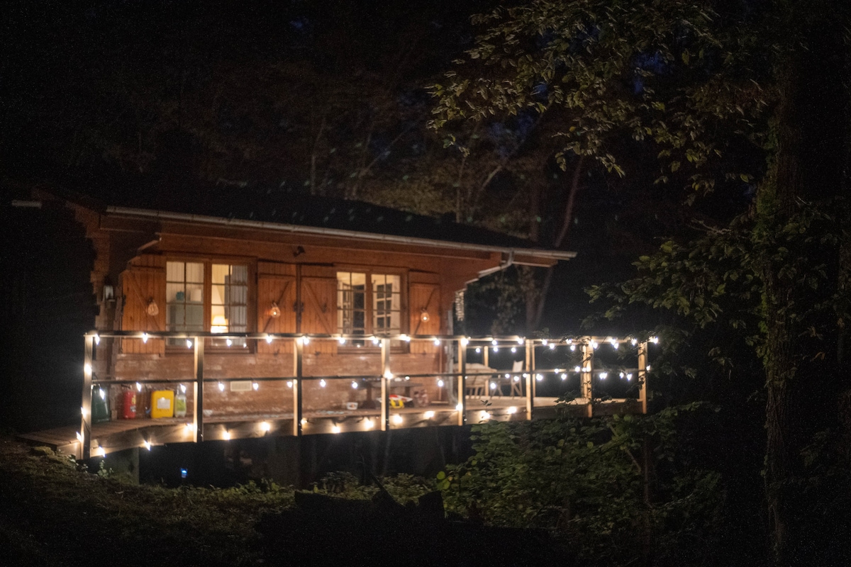 The exterior of the wooden chalet is illuminated at night by soft string lights, enhancing the warm glow of its facade. A spacious deck surrounds the entrance, blending seamlessly with the surrounding woodland. Large windows allow glimpses of the inviting interior.