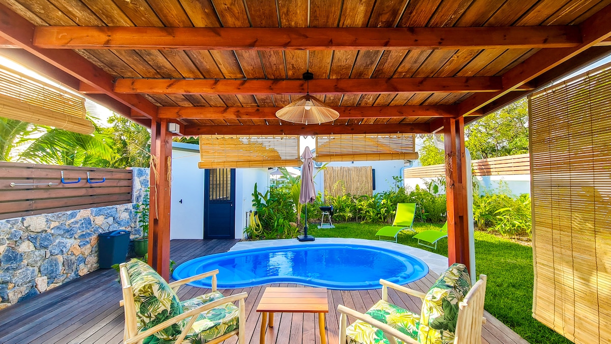 A covered terrace provides shade beside a private swimming pool. Two chairs with tropical-patterned cushions are arranged around a small table. Lush greenery surrounds the area, with a single lounge chair visible in the background. Natural light filters through the bamboo shades.