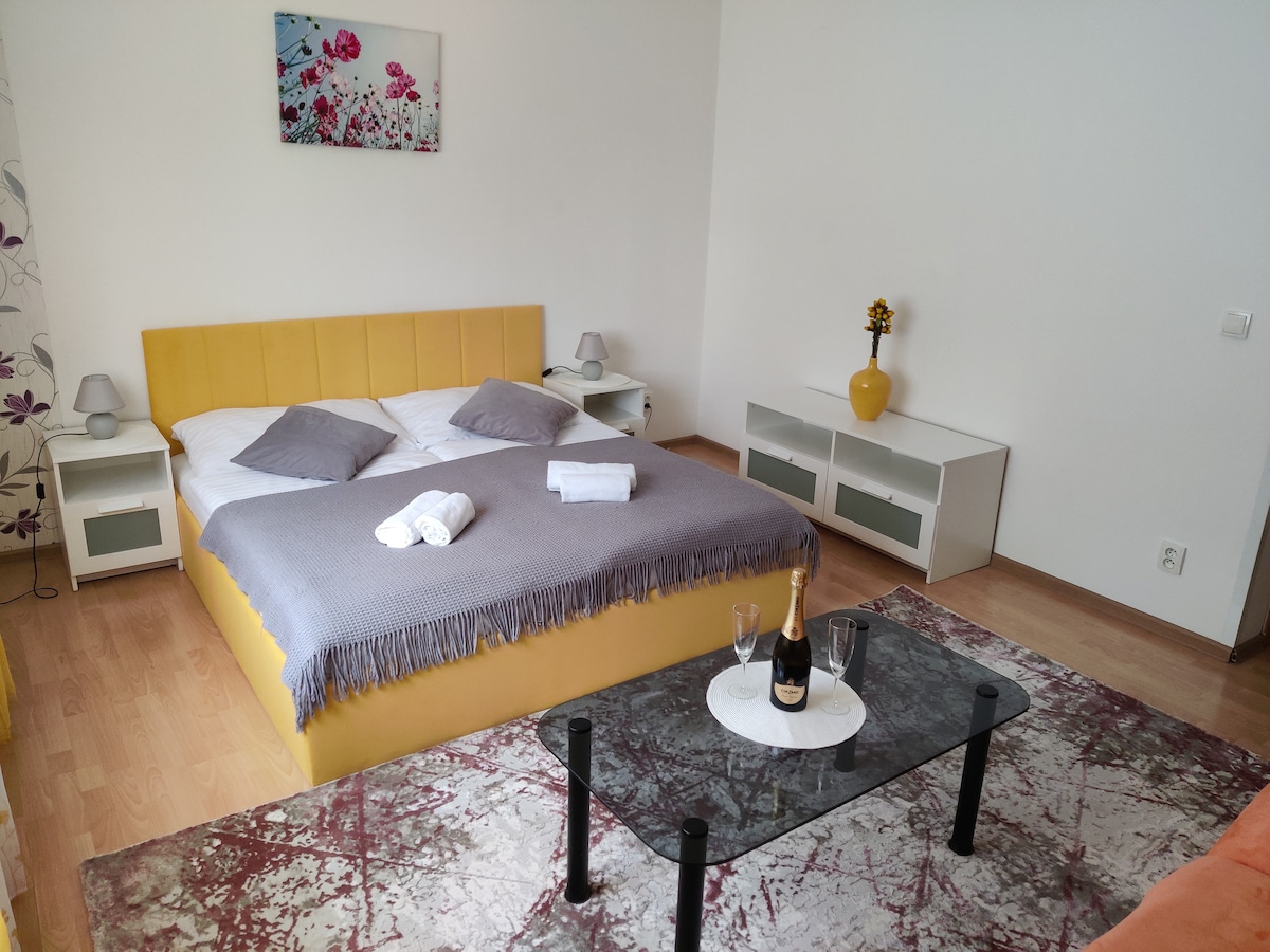 A well-lit sleeping area features a spacious yellow bed with white linens and gray accent pillows. A complementary gray throw is draped over the bed. A glass coffee table is placed in front of the bed, accompanied by two glasses and a bottle of champagne. Decorative elements include a vase with flowers.