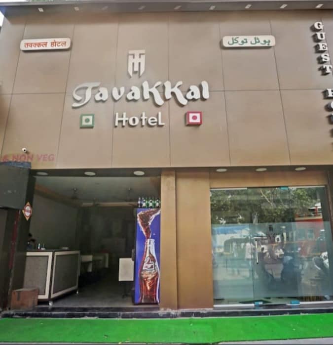 The exterior of a modern hotel is displayed, featuring a streamlined facade with the name 'TavaKKal Hotel' prominently shown. Green grass decorates the entrance, and large windows provide a glimpse of the interior. Signs are visible in multiple languages, indicating the establishment's guest services.