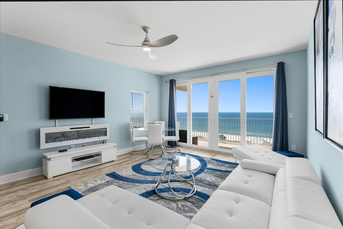 A modern living area features a large sectional sofa in white, complemented by a circular area rug in shades of blue. A flat-screen TV is mounted on the wall. Floor-to-ceiling glass doors lead to a balcony with expansive ocean views.