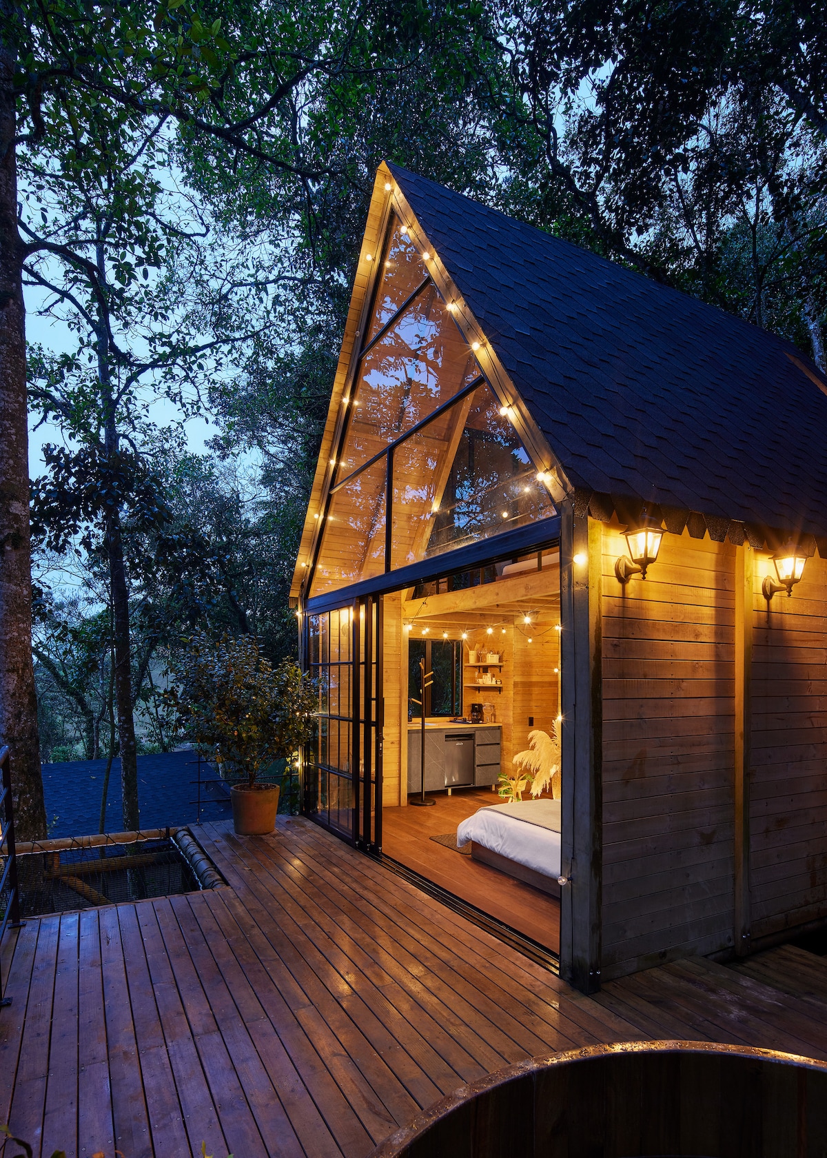 A two-story cabin is set amidst lush trees, featuring a large glass front that opens to a wooden deck. Soft lighting enhances its exterior, while the inviting interior reveals a glimpse of the cozy sleeping area and fully equipped kitchen.