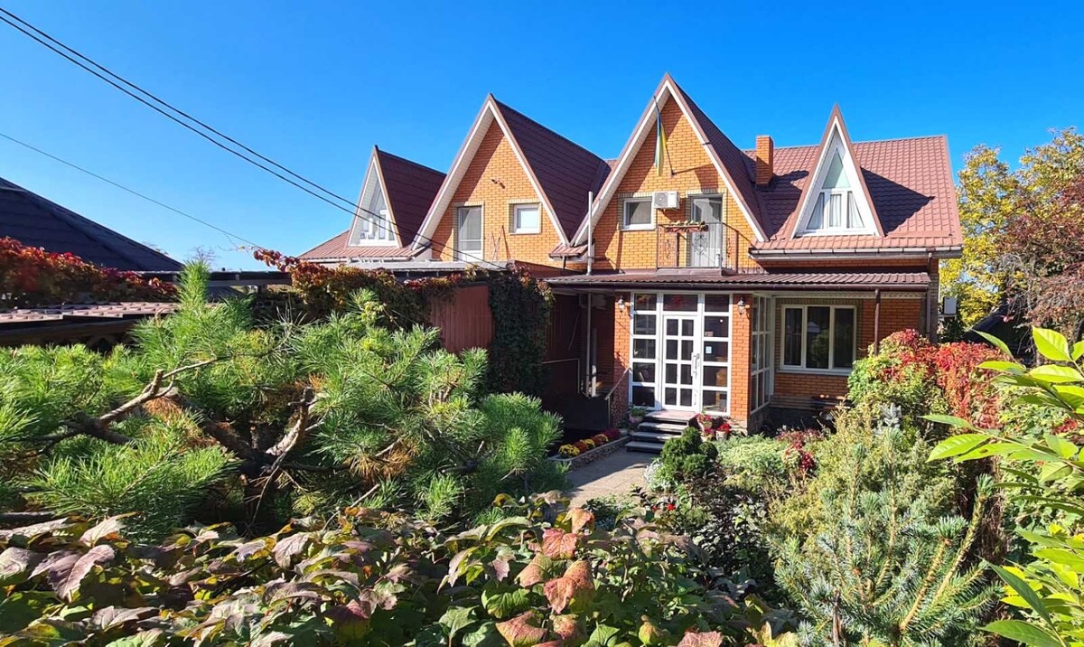 A charming multi-story home is surrounded by lush greenery and colorful foliage. The distinct triangular roofs complement the brick exterior. A welcoming entrance features a large glass door, inviting natural light into the space. A clear blue sky serves as the backdrop.