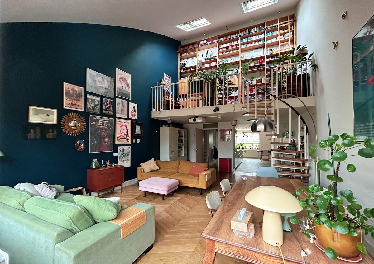 A spacious living area is highlighted by high ceilings and an open layout. The dark blue wall showcases various artworks. A comfortable green sofa, a pink ottoman, and a wooden dining table are visible, along with a staircase leading to a lofted area filled with plants and books.