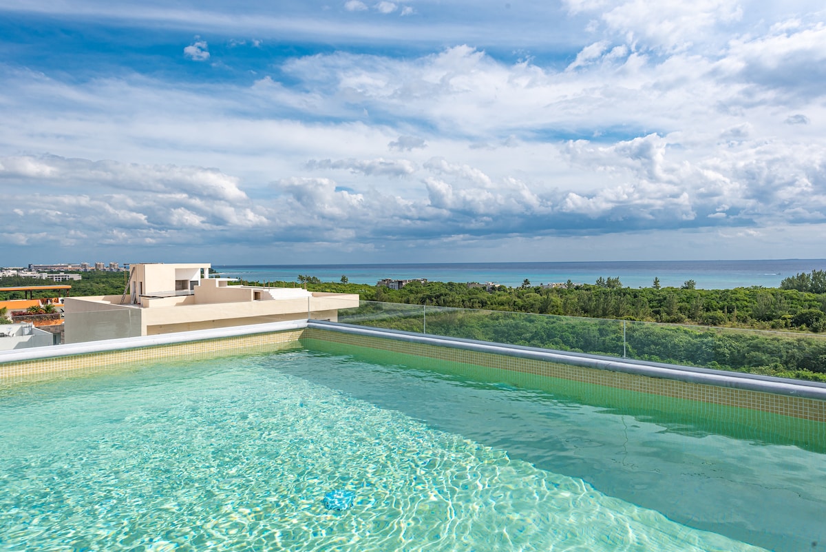 An infinity pool stretches across the rooftop, showcasing tranquil water reflecting the sky. The horizon features a blend of greenery and ocean, under a vast sky filled with soft, wispy clouds.