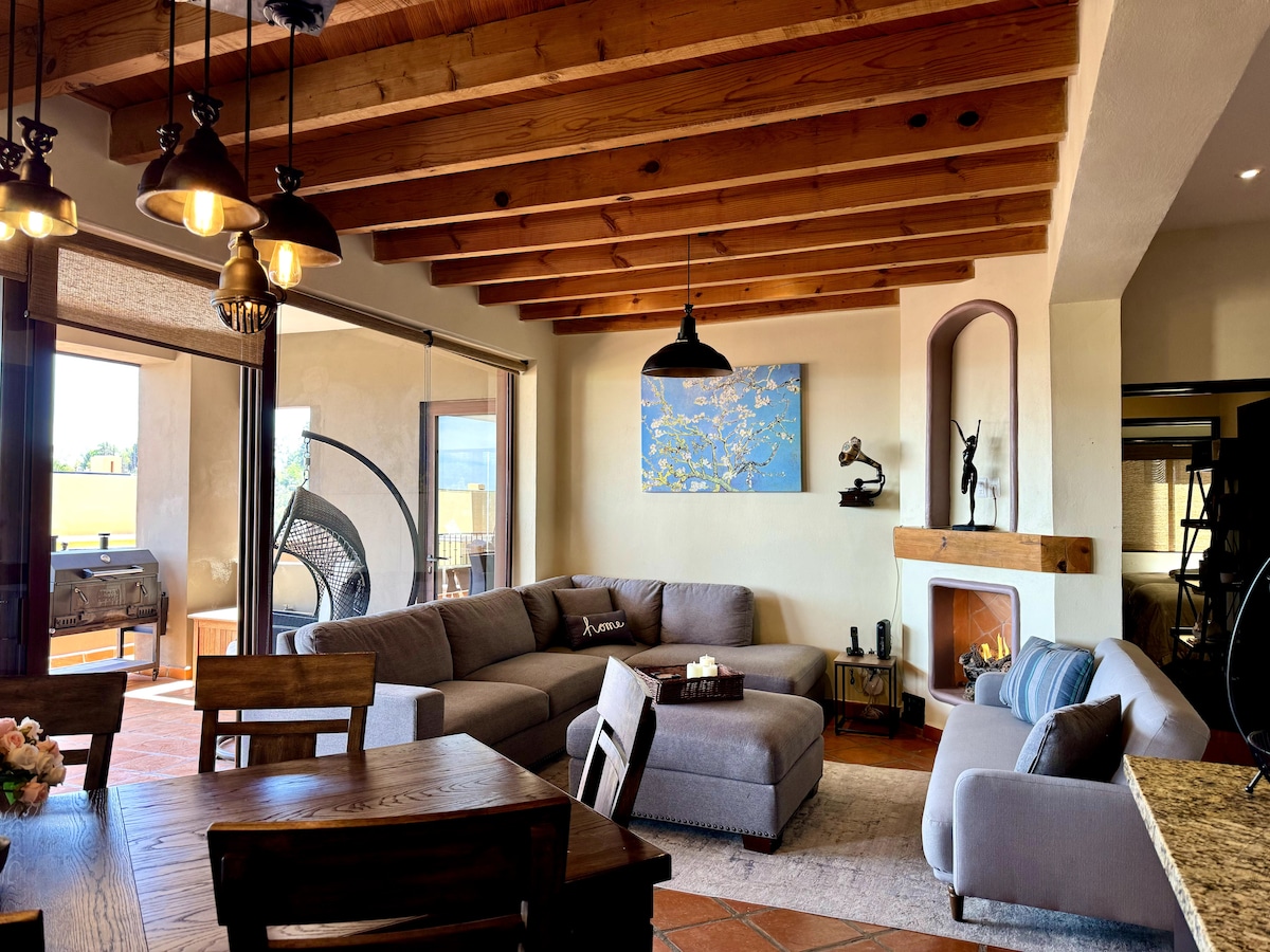 An open-concept living space features a comfortable sectional sofa, a dining area with a wooden table, and a gas fireplace. Large glass doors lead to a spacious terrace. Wood beams adorn the ceiling, while a piece of art adds a touch of color to the wall.