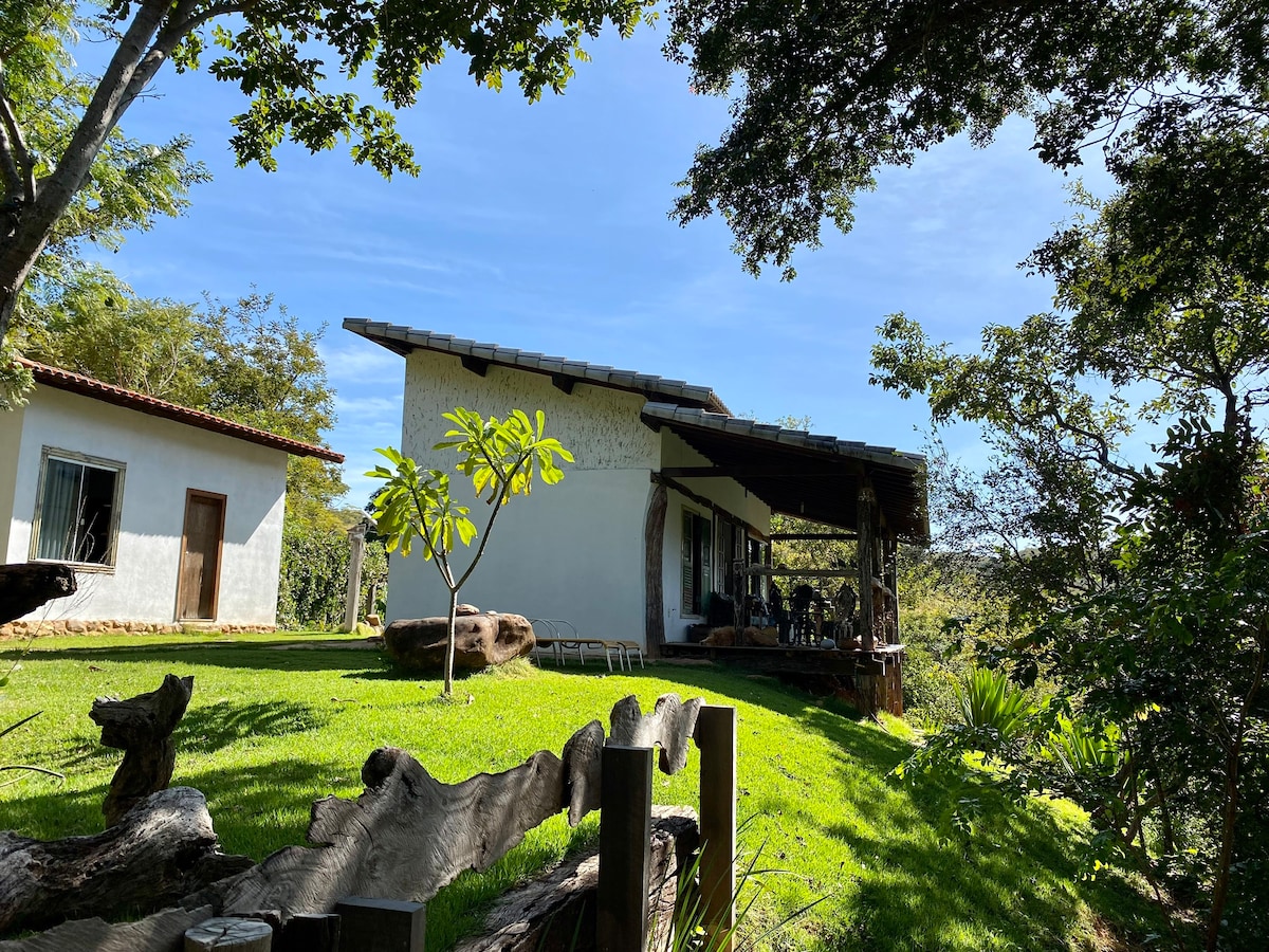 A charming exterior view of a white cottage is highlighted by lush greenery and trees. The structure features a covered porch supported by wooden beams, while comfortable seating is arranged on the porch, surrounded by a vibrant yard.