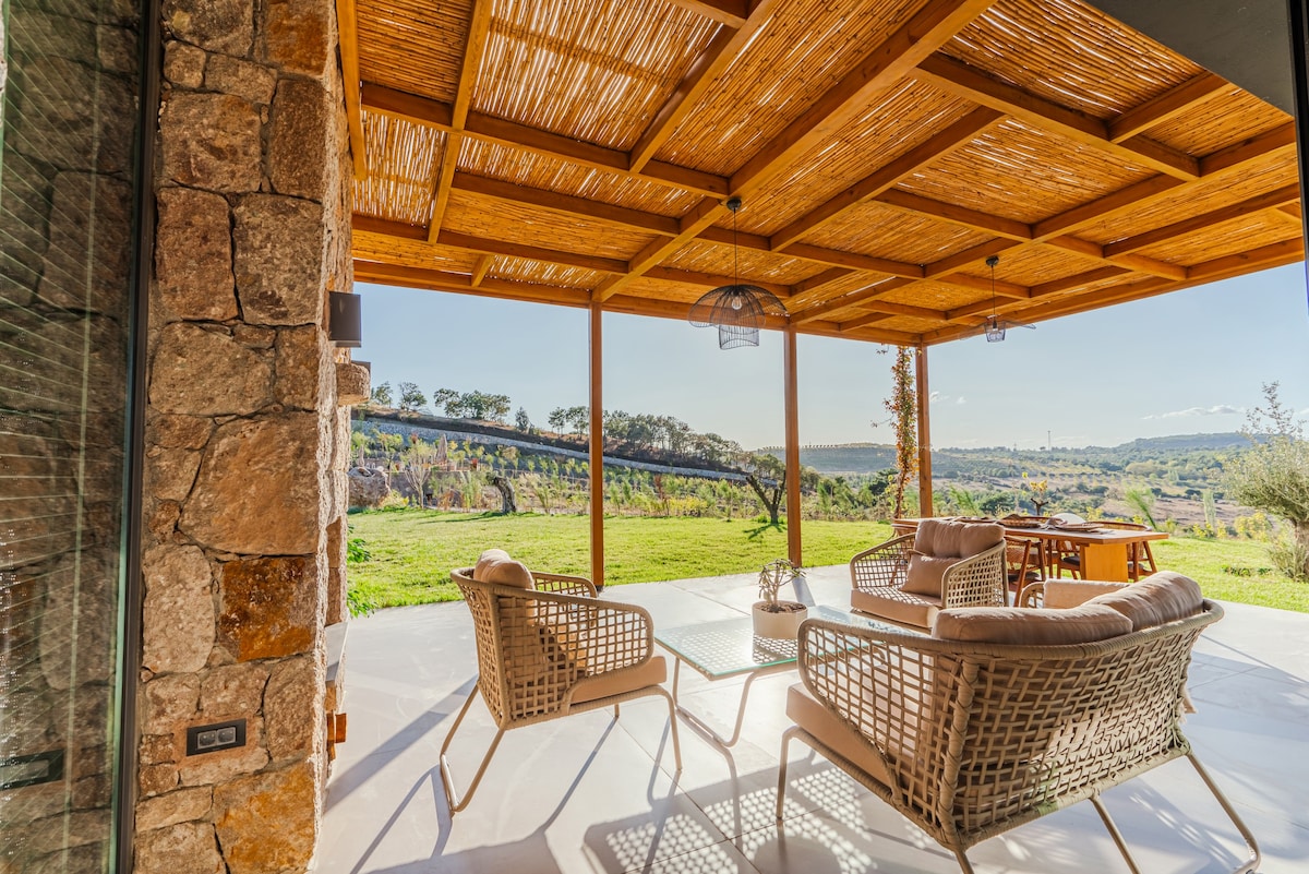 An outdoor seating area is framed by a wooden pergola, providing shade over wicker chairs and a small table. Lush green fields extend into the distance, creating a serene backdrop. Natural light floods the space, enhancing the open and inviting atmosphere.